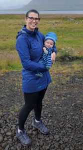 A woman smiles in a rain coat and running attire holding a baby boy with a blue hat on with a lake and mountains in the background
