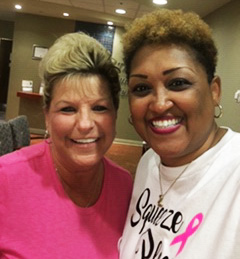 A woman in pink scrubs hugs and smiles with a woman in a white pink ribbon t-shirt