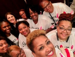 Ten women smiling doing a selfie and wearing white t-shirts that say "Squeeze Pleez" with pink ribbons