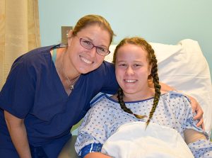 Female Doctor smiling in glasses and blue scrubs with arm around smiling patient with braids and gown