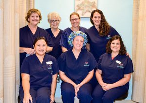 Seven women in dark blue scrubs smile in front of a light blue wall