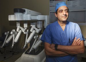 VPFW's Dr. Ramzi Aboujaoude, urogynecologist wearing blue scrubssmiles with arms crossed in front of robotics surgery equipment for Chesterfield Observer