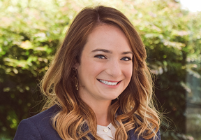 Dr. Kristen Lawson brunette smiling with white top and navy coat in front greenery