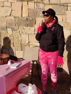 Woman in pink gloves, pants, and pink ribbon jacket speaking into a pink microphone