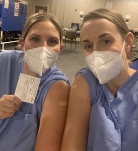 Dr. Schnatz and Dr. Hempel, smiling under their masks, showing their band-aided arms and COVID-19 vaccine cards