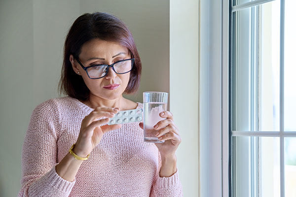 Middle aged woman with glasses looking at menopause hormone therapy pills and holding a glass of water