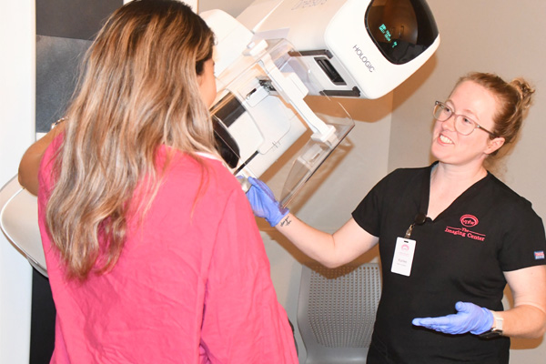Mammography tech in black scrubs with pink VPFW logo giving a mammogram and talking to patient