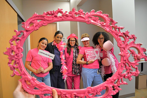 VPFW doctors and nurses wearing pink boas, hats, scarves and boxing gloves and breast cancer awareness signs, viewed through a pink ornate frame.