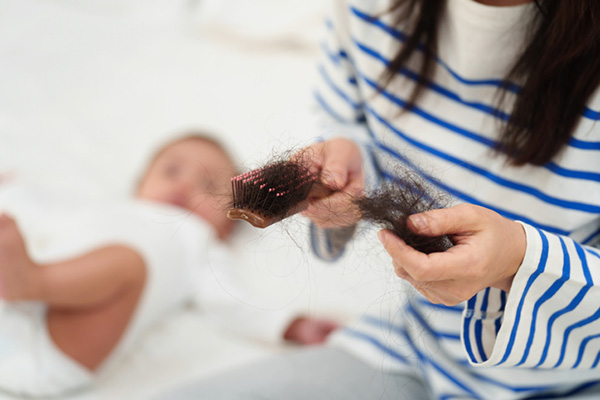 A postpartum woman examines the hair stuck in her hairbrush while her baby lays nearby