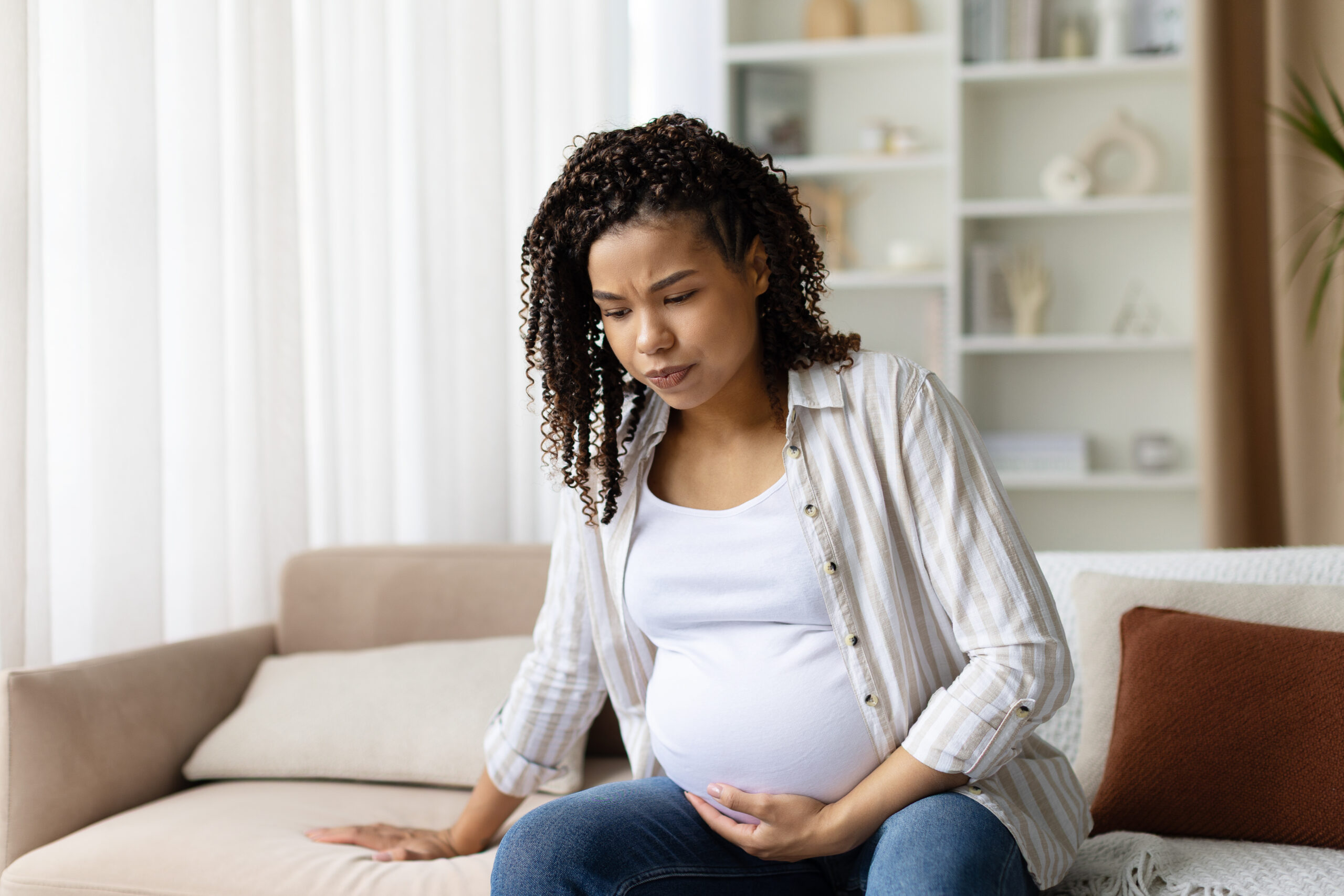 A pregnant woman sits on the sofa, holding her belly in contemplation
